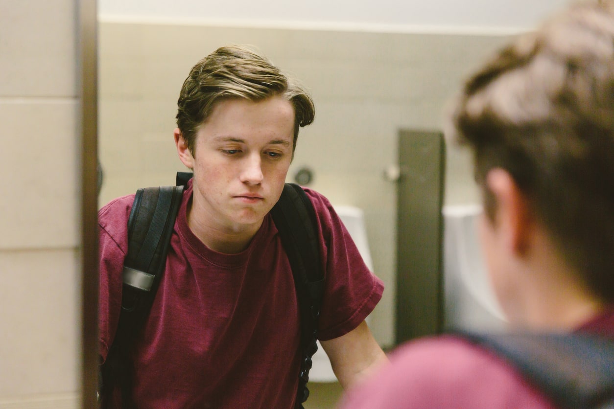 Depressed teen boy looks in the bathroom mirror at school