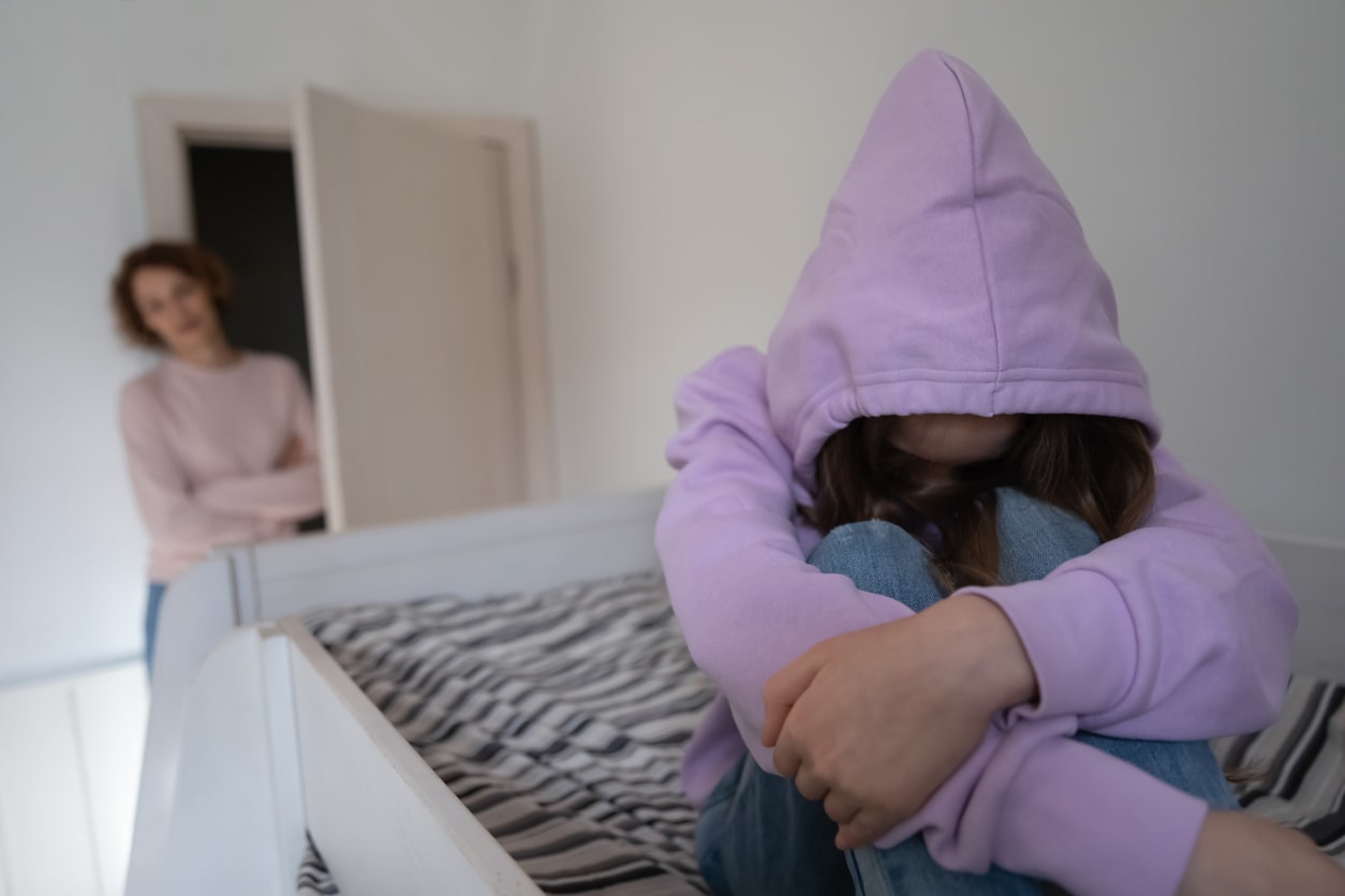 Depressed teen girl wearing hood sitting on bed ignoring step mother